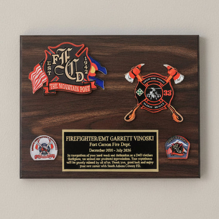 A rectangular wooden firefighter memorial plaque with a dark brown wood grain finish, featuring a gold-toned inscription plate and several firefighter-themed emblems and badges, photographed from above on a light marble surface.