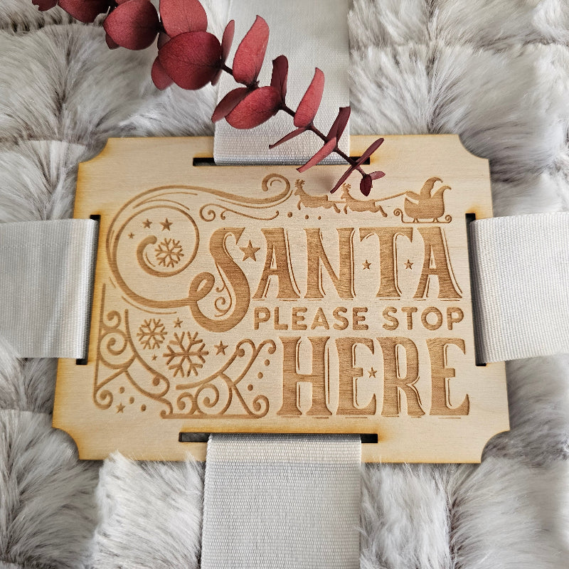 Decorative wooden sign with 'Santa, please stop here' text on a fluffy white surface.