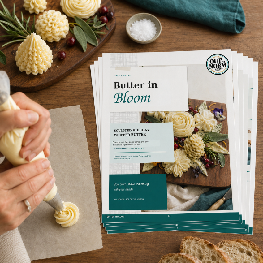A person's hands are shown piping cream-colored butter onto parchment paper, with a stack of printed recipe cards titled 'Butter in Bloom' on the table. The scene is styled with butter sculptures, bread slices, a small bowl of salt, and a teal cloth on a wooden surface.