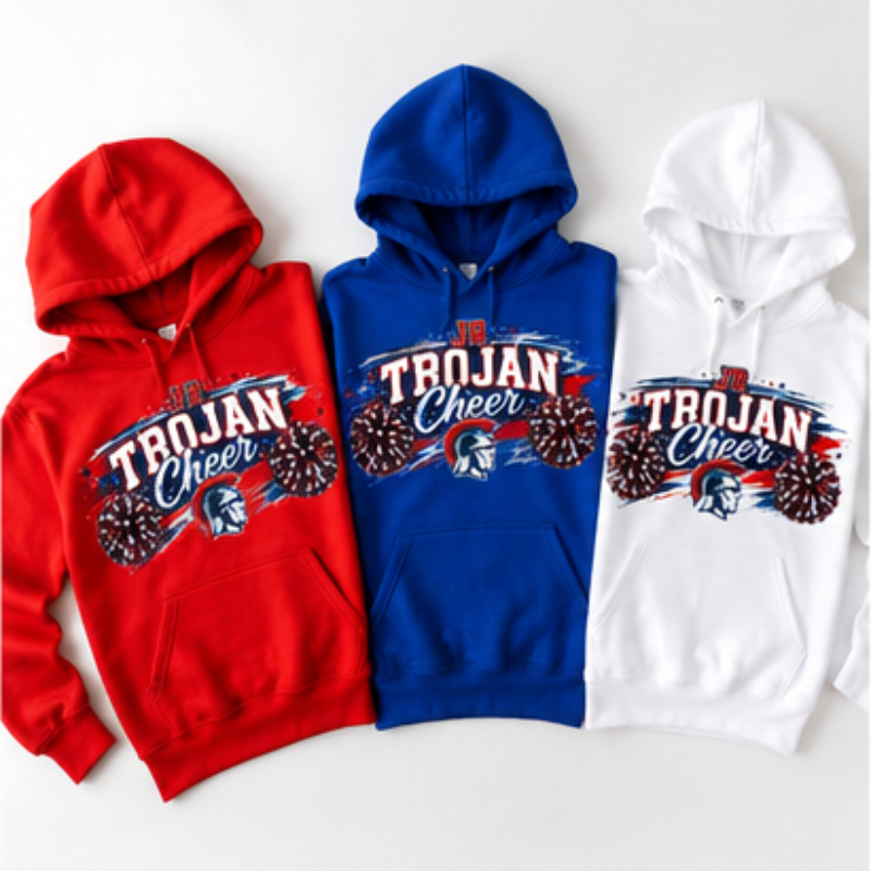 Three hooded sweatshirts shown flat against a light gray background: a red hoodie, a royal blue hoodie, and a white hoodie. Each has a large front graphic reading 'TROJAN Cheer' with a Spartan helmet and fireworks motif in red, white, and blue.