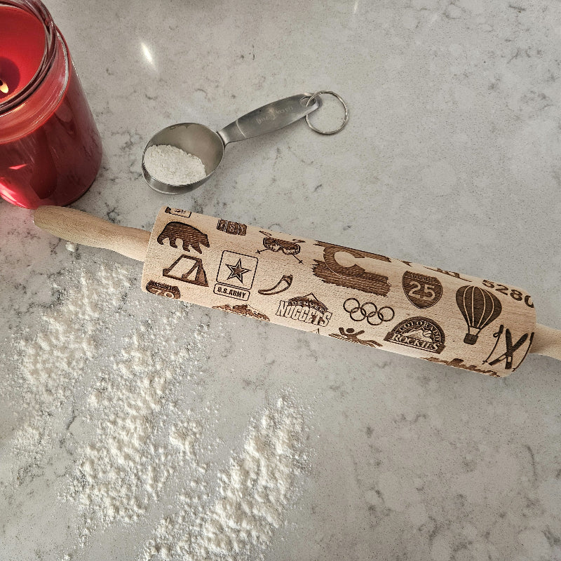 Rolling pin with embossed designs on a light surface next to a lit red candle and measuring spoons.