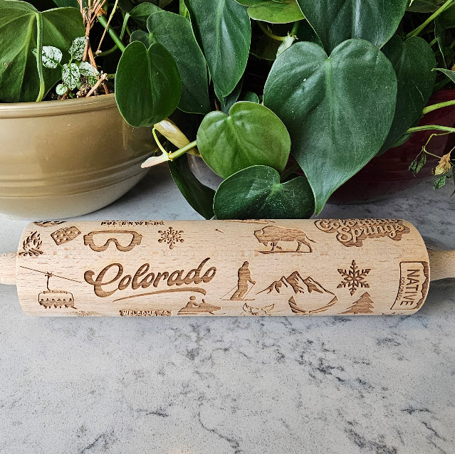 Wooden rolling pin with engraved designs and 'Colorado' text on a marble surface with plants in the background.