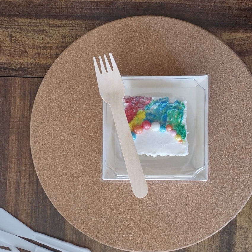 Plastic fork on a round paper placemat with a small square dish containing colorful items on a wooden table.