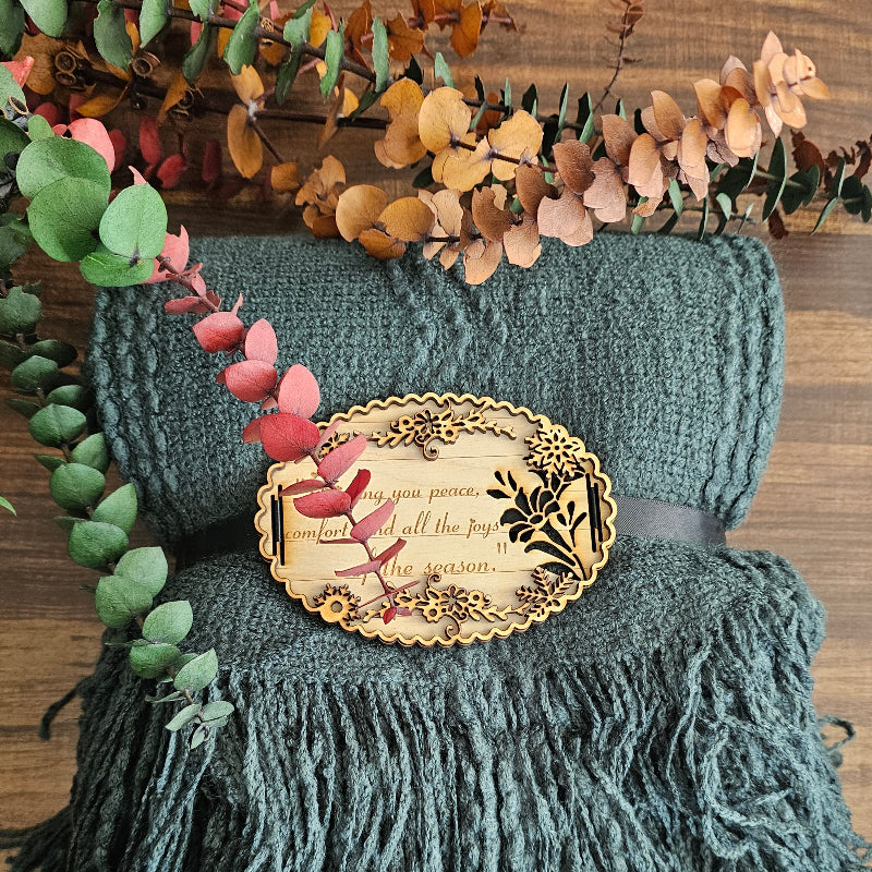 Teal blanket with fringe and decorative brooch on a wooden surface with foliage.