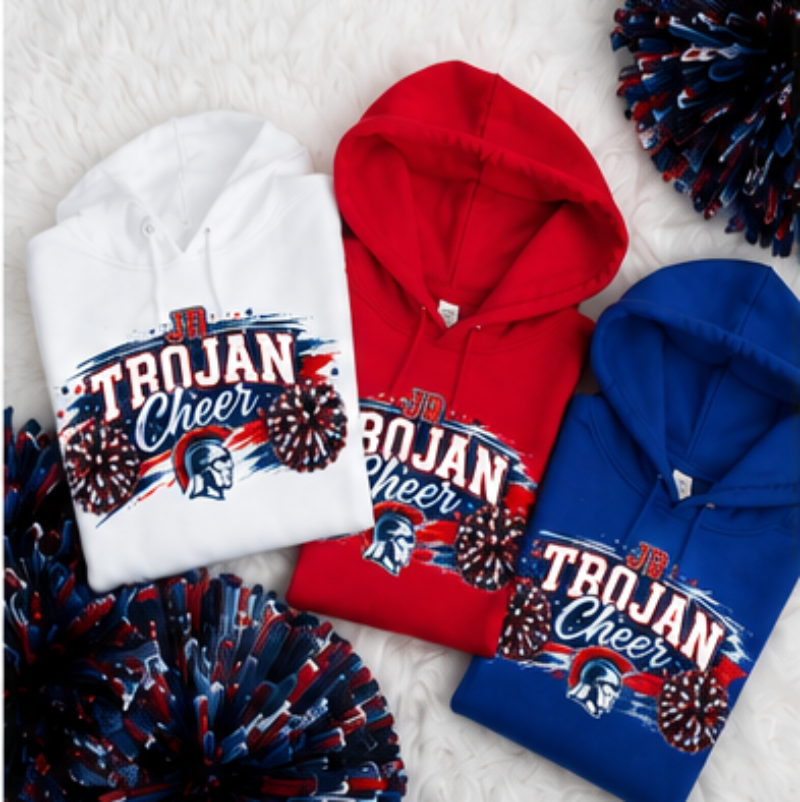Three folded hoodies in white, red, and blue are arranged on a white faux-fur surface with patriotic pom-poms. Each hoodie has a large front graphic reading 'TROJAN Cheer' with a Spartan helmet and fireworks motif.