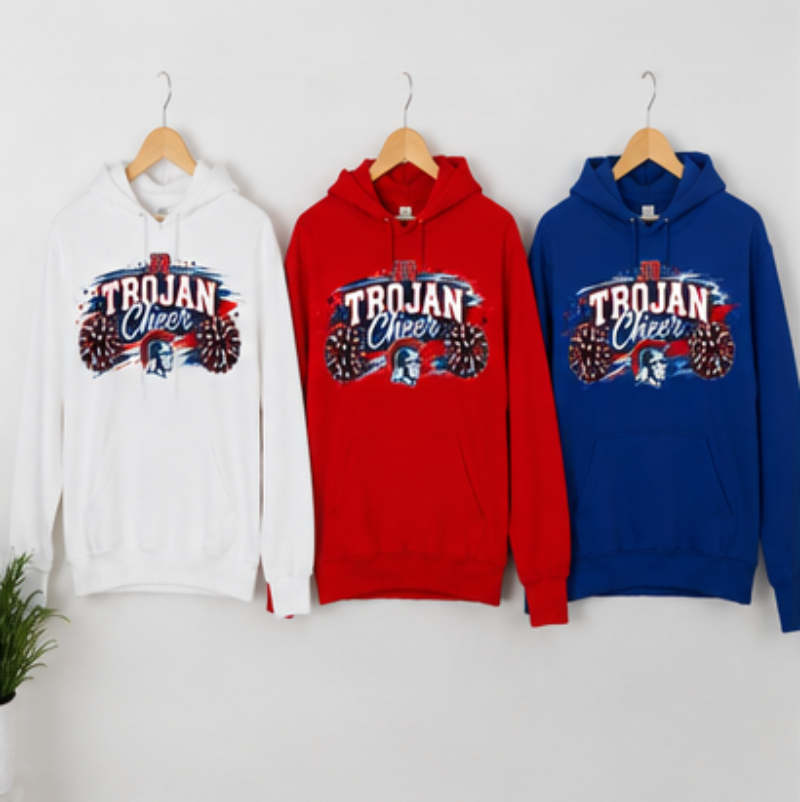 Three hoodies hanging on wooden hangers against a light gray wall: a white hoodie, a red hoodie, and a blue hoodie, each with a large front graphic reading "TROJAN Cheer" and a football-themed design.
