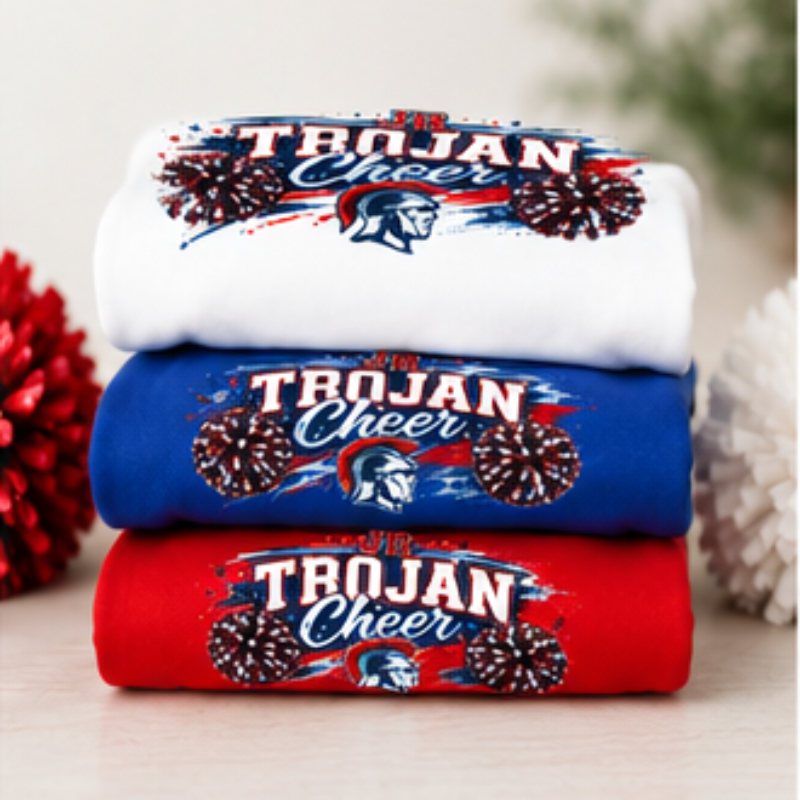 Three folded T-shirts stacked vertically in white, blue, and red, each printed with 'TROJAN Cheer' and a Spartan helmet graphic, set against a softly blurred holiday background.