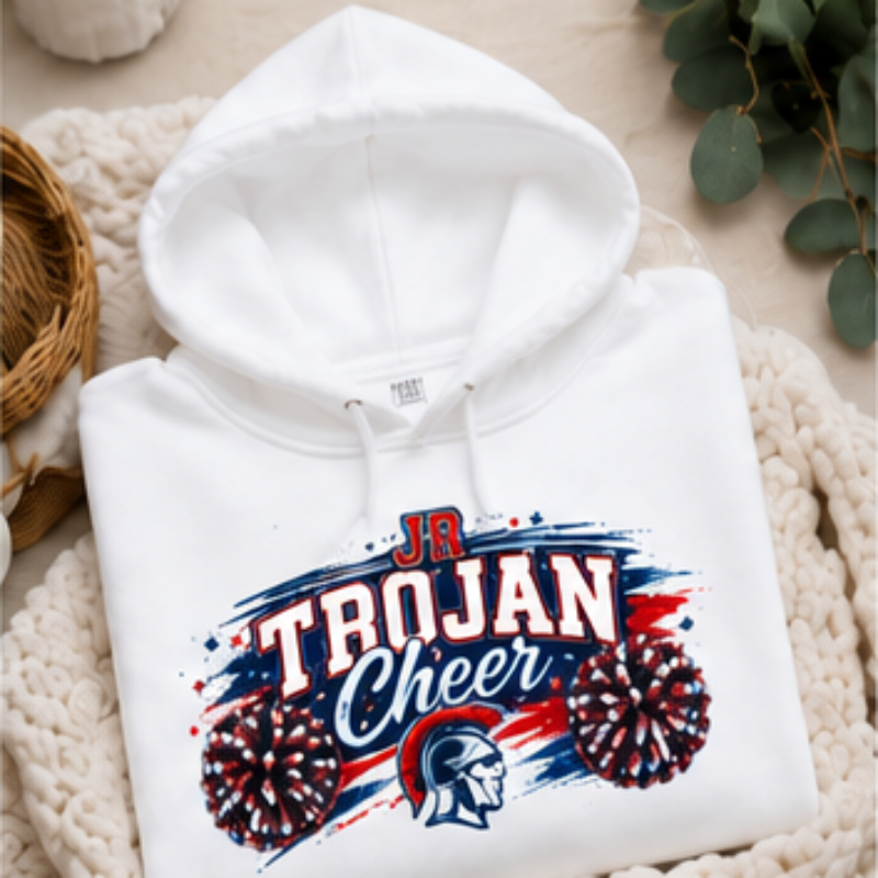 A folded white hoodie with a 'JR TROJAN Cheer' graphic in red, white, and blue, styled on a chunky cream knit blanket with decorative eucalyptus leaves.