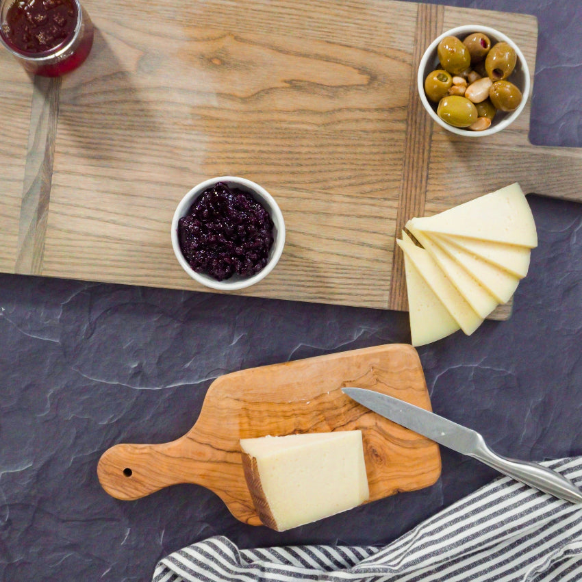 Cheese and jam platter with cutting boards, knives, and a striped cloth on a dark surface.