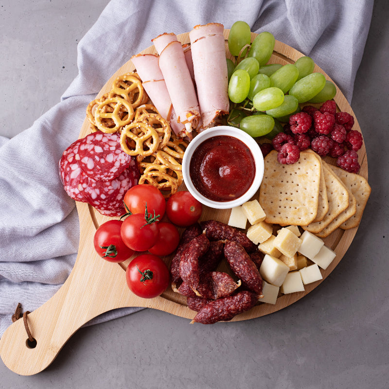 Wooden platter with assorted meats, cheeses, fruits, and crackers on a gray surface.