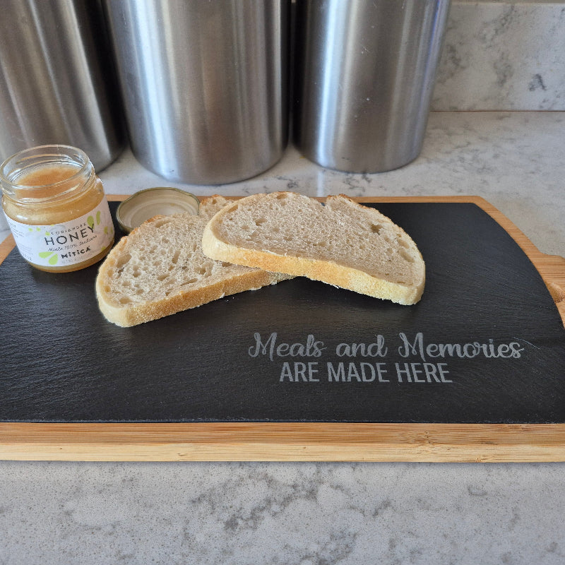 Black slate cutting board with wooden edges displaying 'Meals and Memories ARE MADE HERE' text, accompanied by bread and honey on a kitchen counter.