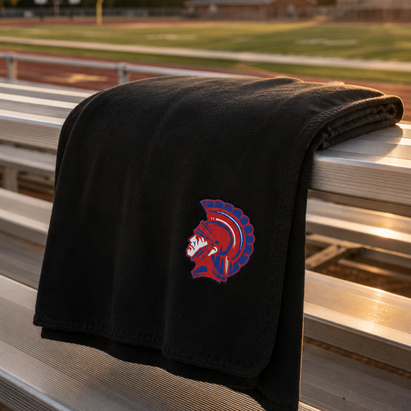 A black fleece blanket with a red and blue Spartan helmet logo, draped over a metal bleacher at a sports field during sunset.