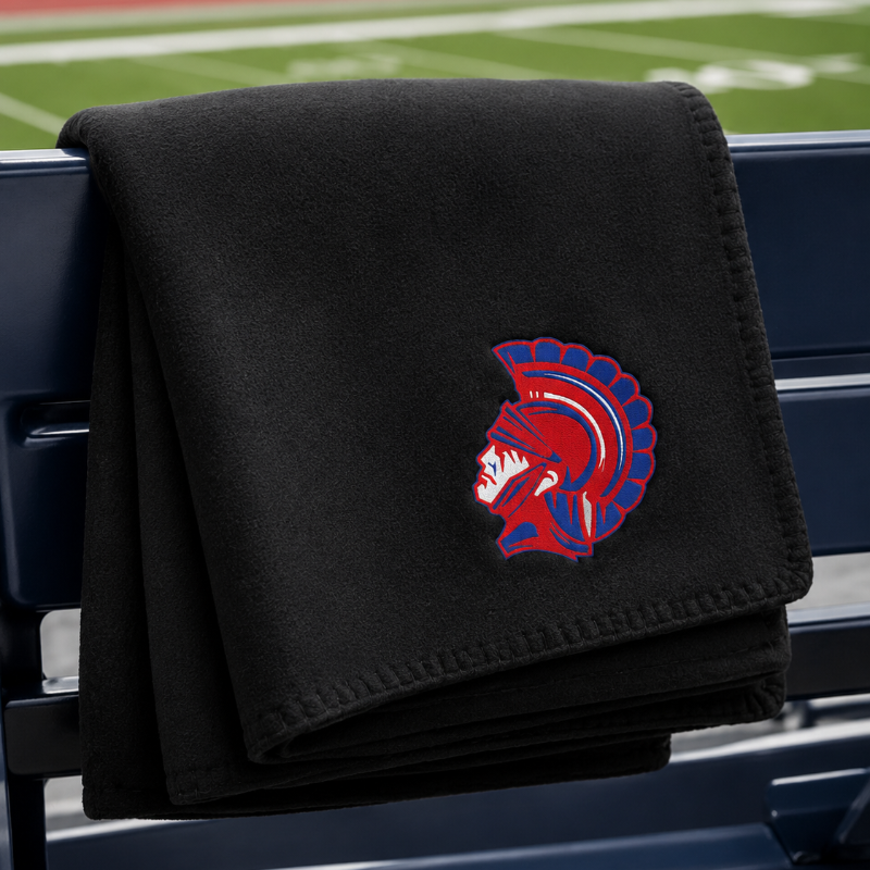 A black fleece throw blanket draped over a blue stadium bench, featuring a red and blue Spartan helmet logo embroidered on the front. The background shows a blurred football field and track.