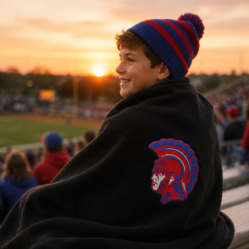 A young boy wrapped in a black fleece blanket sits on stadium bleachers at sunset, wearing a blue and red striped knit beanie. The blanket features a red and blue Spartan helmet logo on the back.