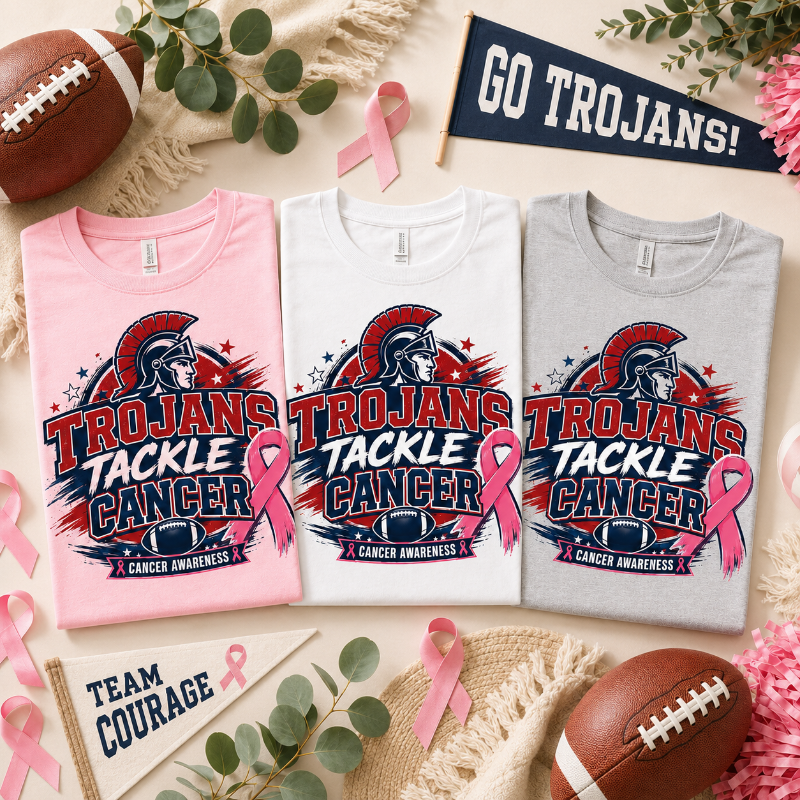 Three folded T-shirts in pink, white, and heather gray are arranged in a flat lay with footballs, pink ribbons, eucalyptus leaves, and small signs that read GO TROJANS!, TEAM COURAGE, and TEAM TROJANS. Each shirt features a large Trojan helmet graphic and the words 'TROJANS TACKLE CANCER' with 'CANCER AWARENESS' below.
