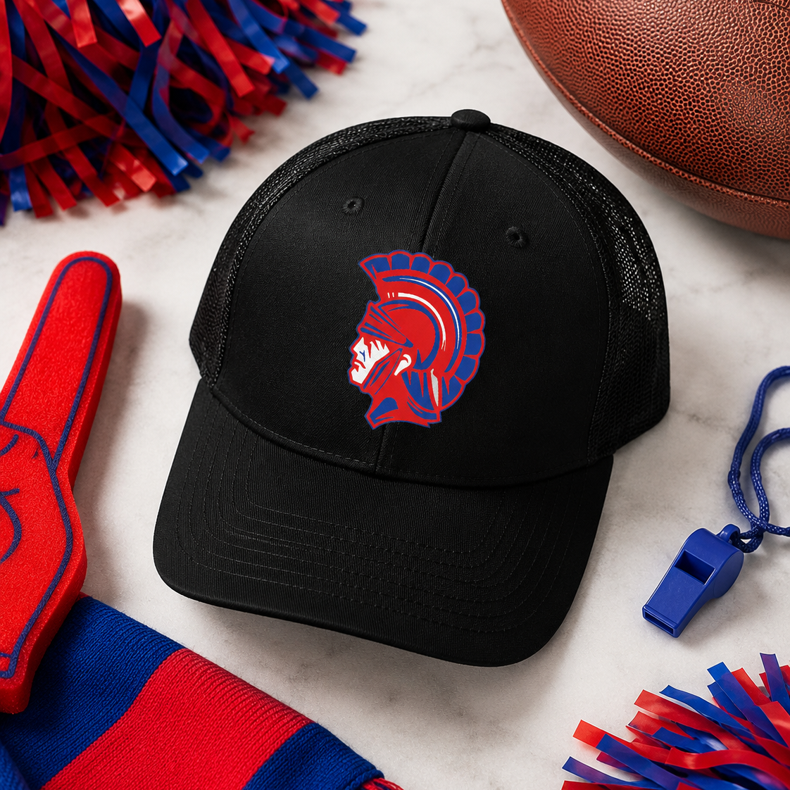A black trucker-style baseball cap with a red and blue Spartan helmet logo on the front, styled on a white marble surface with football, pom-poms, a foam finger, a scarf, and a whistle around it.