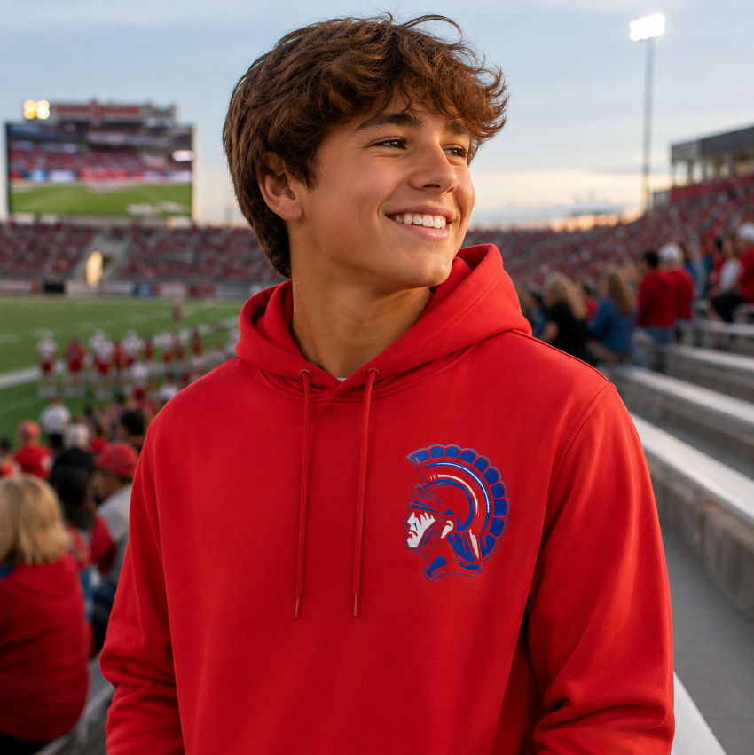 A smiling young man in a stadium stands wearing a bright red pullover hoodie with a blue and white Spartan helmet logo on the left chest. The background shows a football field, stadium seating, and other spectators.