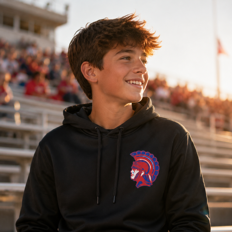 A smiling young man sitting in stadium bleachers at sunset, wearing a black pullover hoodie with a red and blue Spartan helmet logo on the left chest.