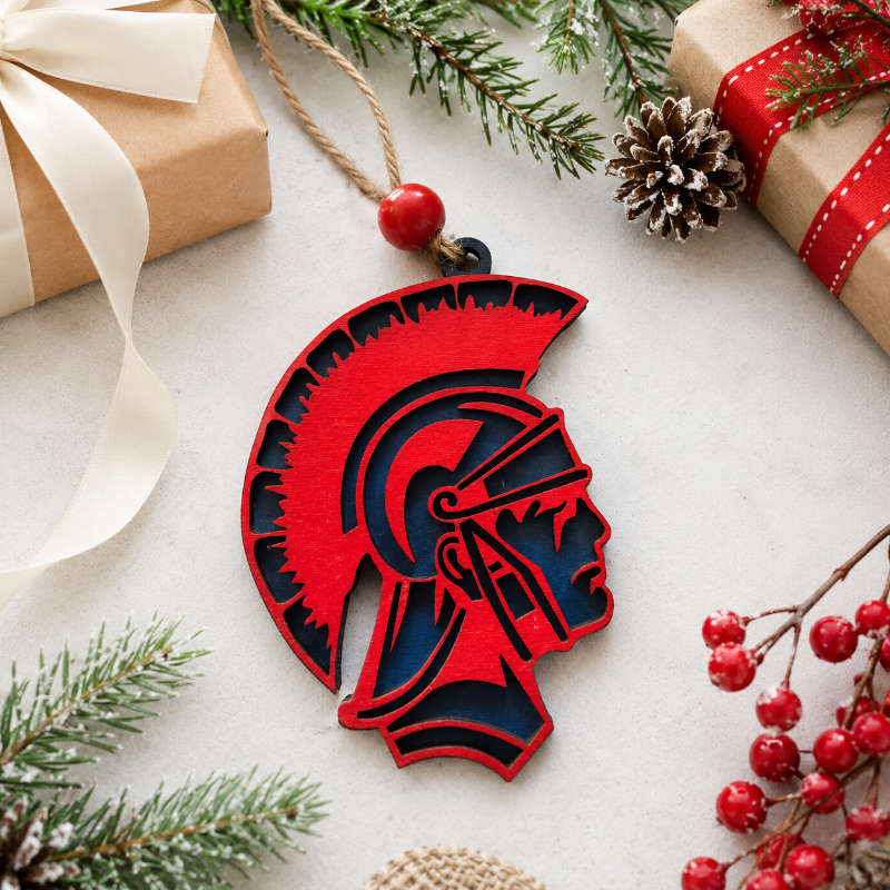 A red and navy blue wooden Spartan helmet Christmas ornament hanging from a twine string with a red bead, styled on a light gray surface with holiday greenery, pinecones, and wrapped gift boxes.