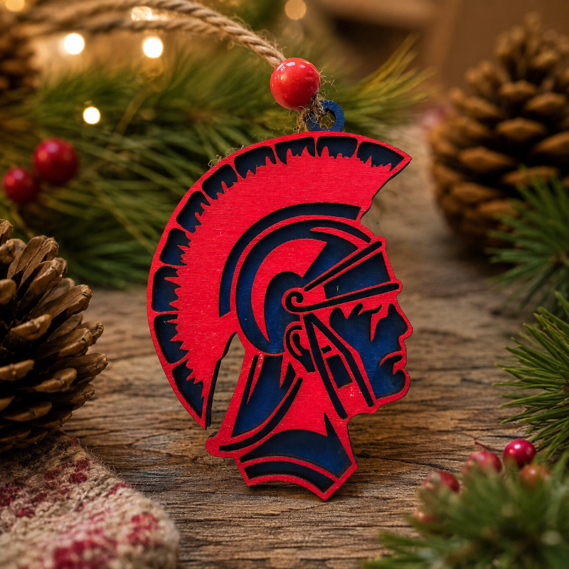A red and navy blue wooden Spartan helmet ornament hanging from a twine string with a red bead, set against a rustic holiday background of pine branches, pinecones, and warm string lights.