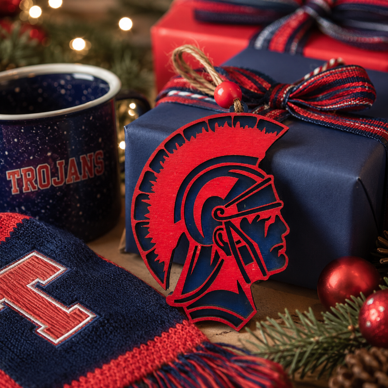 A festive holiday gift scene featuring a red and navy blue Spartan helmet ornament in front of wrapped presents, a navy blue mug with 'TROJANS' text, and a matching knit scarf with pine branches and ornaments.