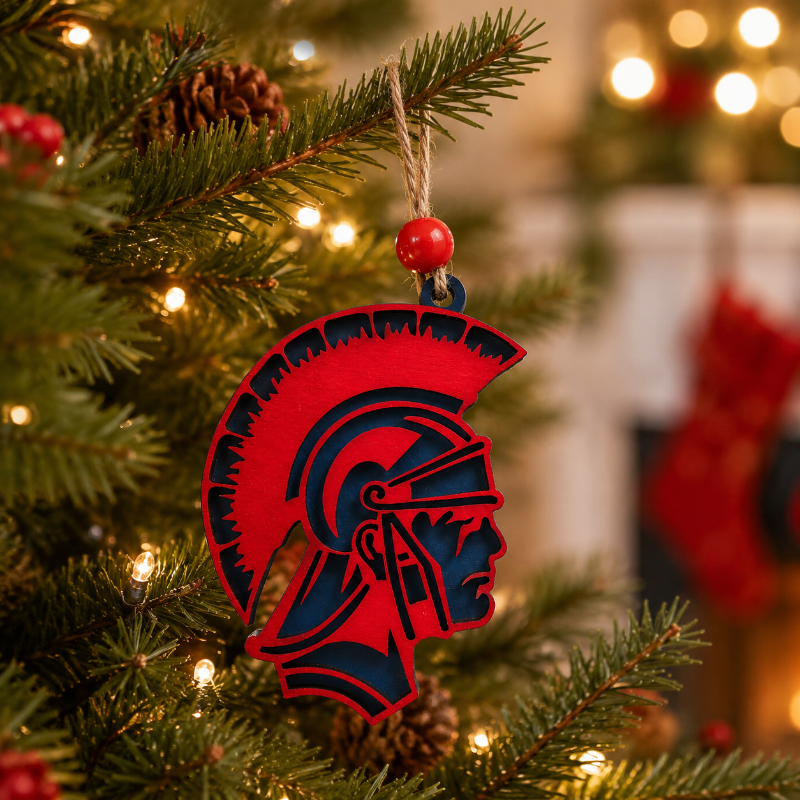 A red and navy blue Spartan helmet Christmas ornament hanging on a decorated Christmas tree with warm string lights.