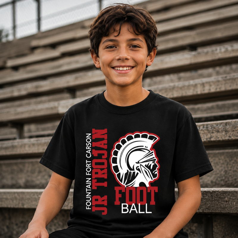 A smiling young boy sits on outdoor concrete stadium steps wearing a black short-sleeve t-shirt with a red, white, and gray graphic that reads 'Fountain Fort Carson Jr. Trojan Football' alongside a Spartan helmet logo.