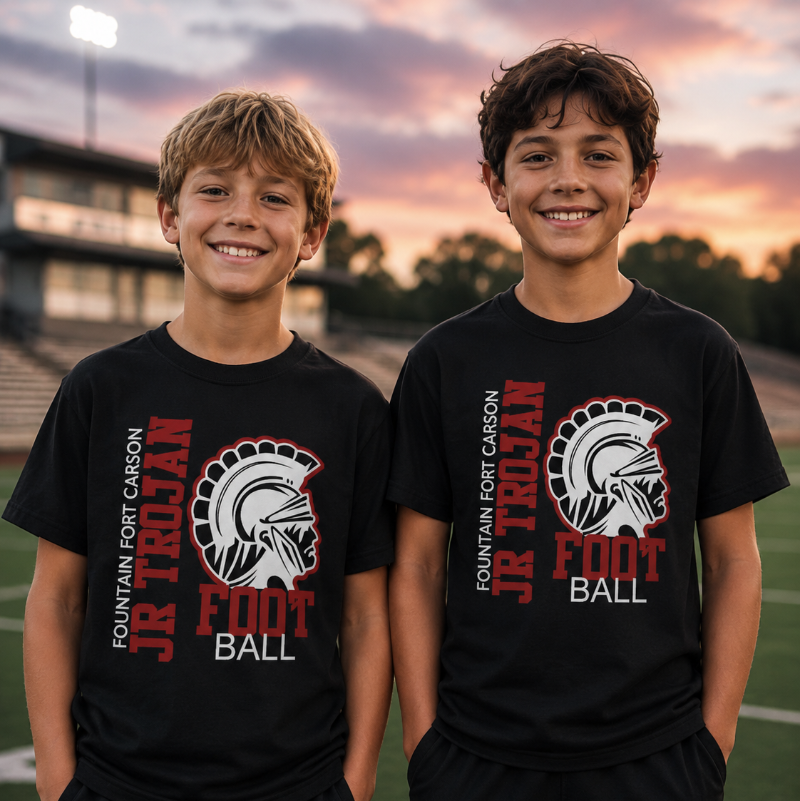 Two boys standing on a football field at sunset wearing matching black t-shirts with a red, white, and gray Spartan helmet graphic and the text 'FOUNTAIN FORT CARSON JR TROJAN FOOT BALL'.