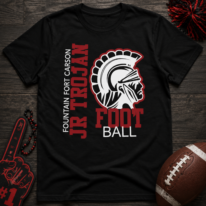 A black short-sleeve t-shirt laid flat on a dark wood surface, featuring a red, white, and black graphic that reads 'Fountain Fort Carson Jr Trojan Football' with a Spartan helmet illustration. A football, pom-poms, and a foam finger are placed around it as styling props.