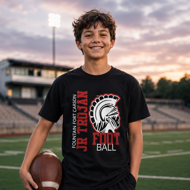 A smiling young man stands on a football field at sunset wearing a black short-sleeve t-shirt with a red, white, and gray 'JR TROJAN FOOTBALL' graphic on the front, paired with black athletic shorts and holding a brown football.