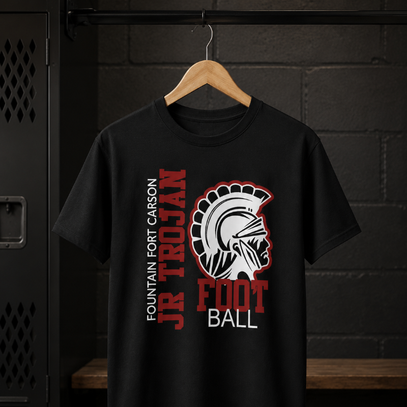 A black short-sleeve t-shirt hanging on a wooden hanger against a dark brick wall and locker. The shirt features a graphic print with the text 'FOUNTAIN FORT CARSON JR TROJAN FOOTBALL' in red and white, alongside the Troyan helmet logo.