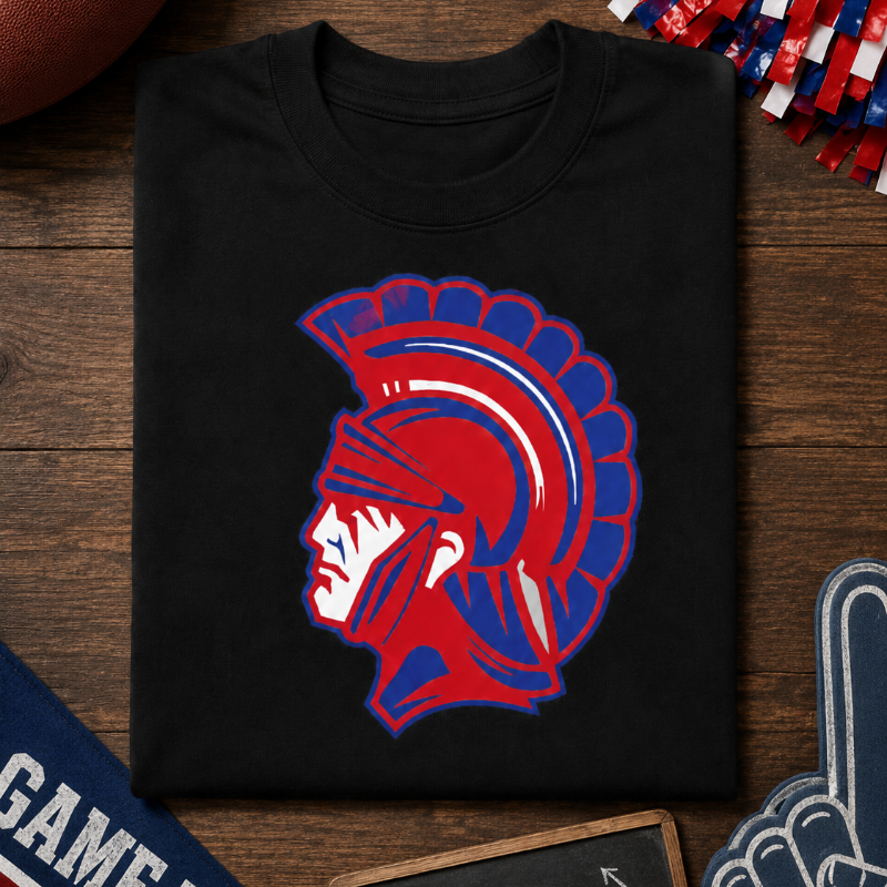 A black crewneck t-shirt featuring a red and blue Spartan helmet graphic, displayed in a flat-lay arrangement on a wooden background with football, pom-poms, and game day accessories.