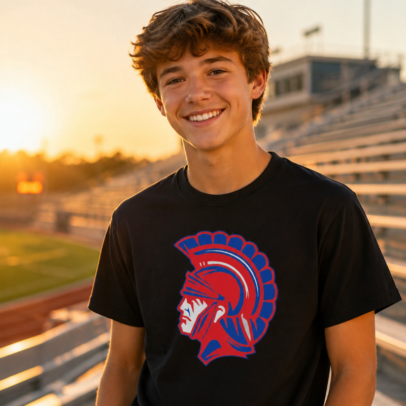 A smiling young man wearing a black t-shirt with a red and blue Spartan helmet graphic, standing on a school track field at sunset.