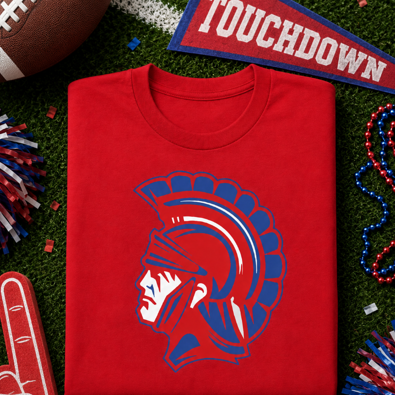A folded red t-shirt with a Spartan helmet graphic in blue and white, displayed on a green turf background with football-themed props like a football, foam finger, and helmet.