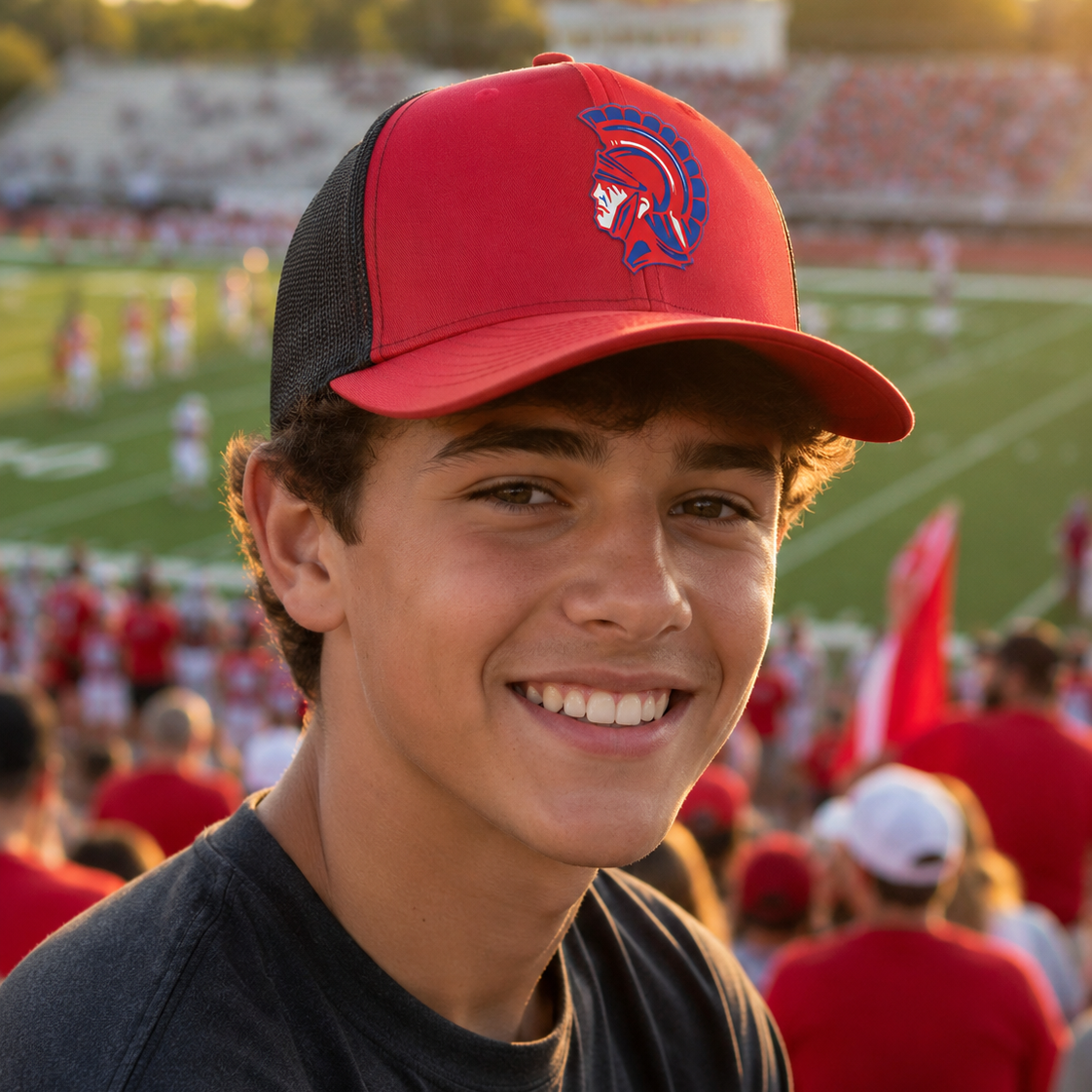 A smiling young man in a football stadium wearing a red and black trucker hat with a Spartan helmet logo, a dark gray t-shirt, and a blurred background of spectators and a field.