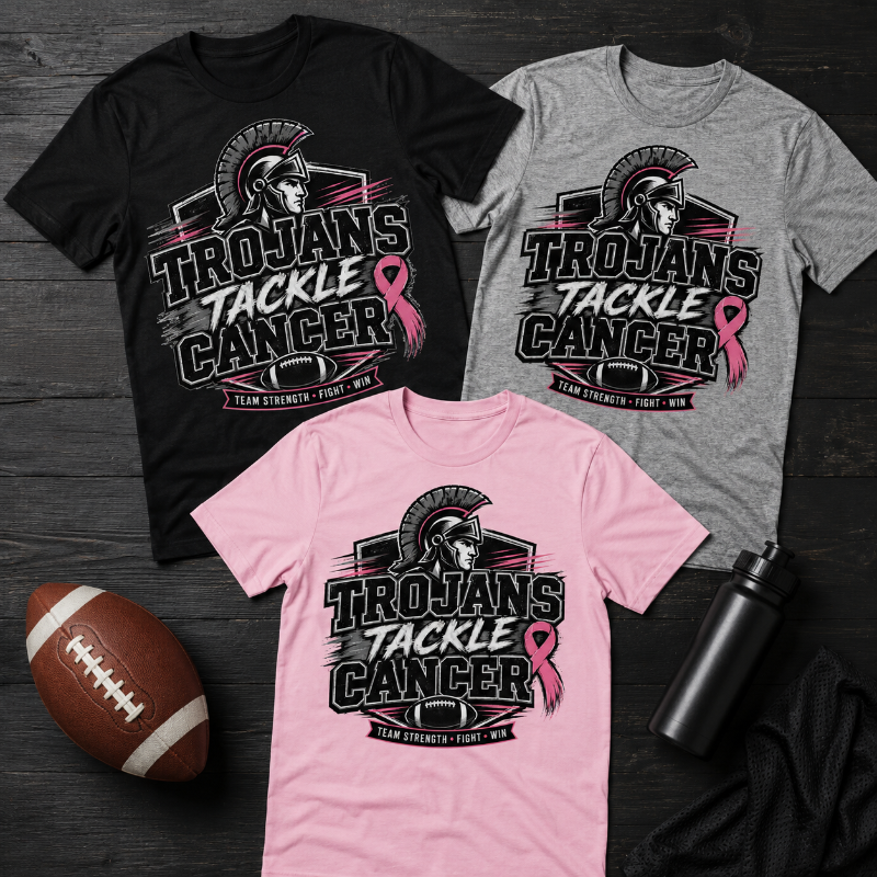Three short-sleeve graphic T-shirts in black, gray, and pink are arranged on a dark wood surface with a football, towel, and water bottle. The shirts feature a large Trojan helmet graphic and the text 'TROJANS TACKLE CANCER' with a pink ribbon and smaller text 'TEAM STRENGTH - FIGHT ON.