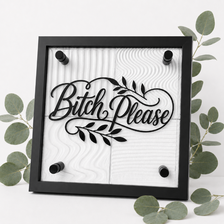 A square black-framed wall sign with the phrase “Bitch Please” in black script over a white wood-grain textured background, styled with eucalyptus leaves on a white surface.