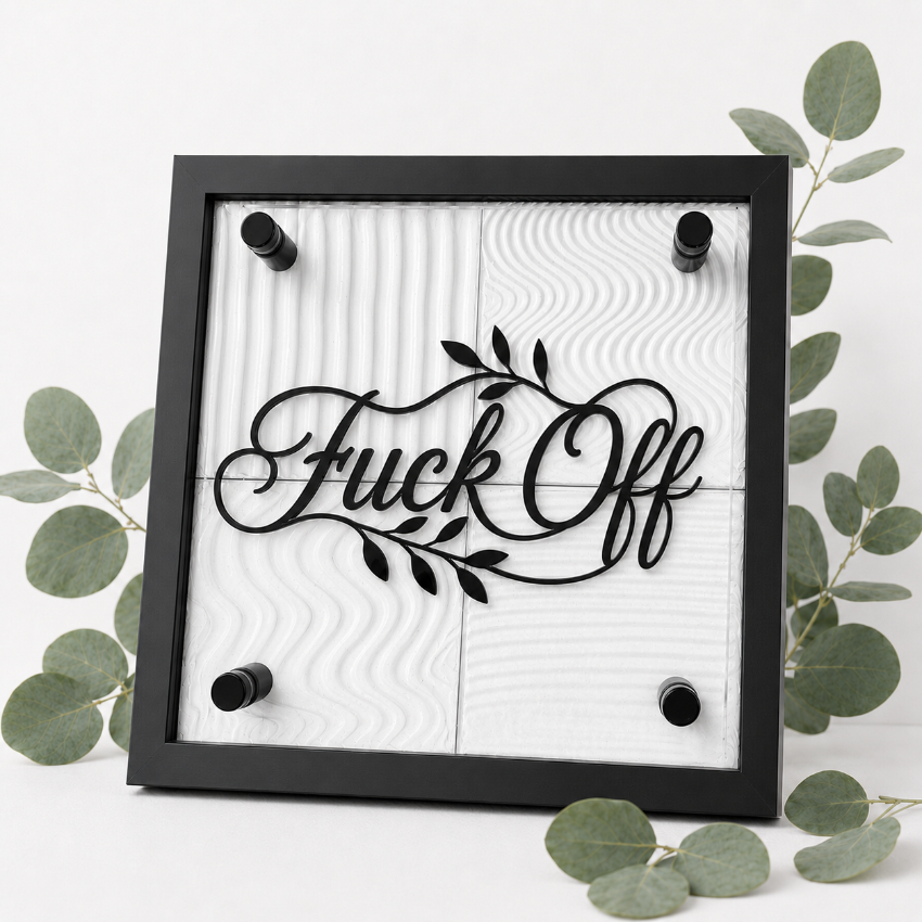 A square black-framed wall sign with the phrase “Fuck Off” in black script over a white wood-grain panel background, surrounded by green eucalyptus leaves on a white surface.