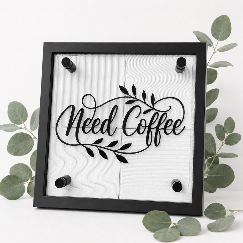 A square framed wall sign with the words “Need Coffee” in black script over a white wood-grain background, surrounded by green eucalyptus leaves on a white surface.