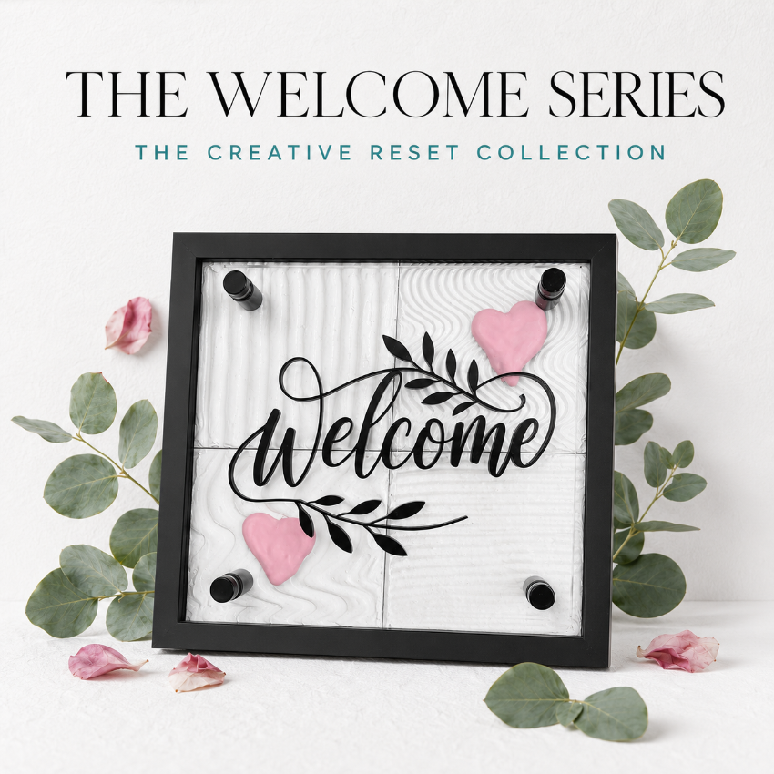 A framed decorative sign reading “Welcome” in black script over a white wood-textured background, with two pink hearts and black mounting dots visible. The sign is styled with eucalyptus leaves and pink petals on a white surface, and the text above reads “THE WELCOME SERIES” and “THE CREATIVE RESET COLLECTION.