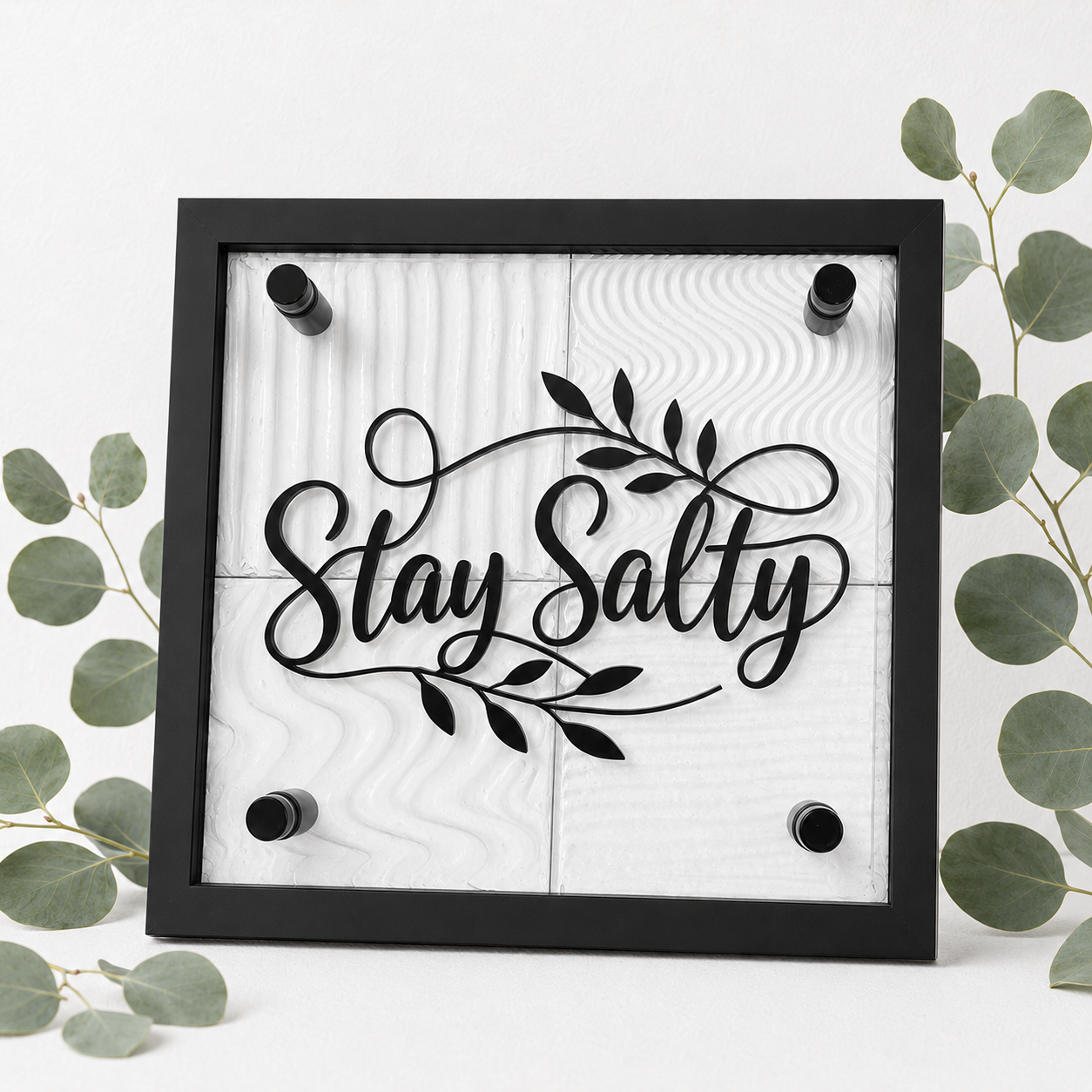 A square black-framed wall sign with the phrase “Stay Salty” in black script over a white wood-grain panel, surrounded by green eucalyptus leaves on a white background.