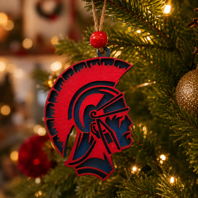 A red and navy blue Spartan helmet Christmas ornament hanging from a tree with warm string lights.