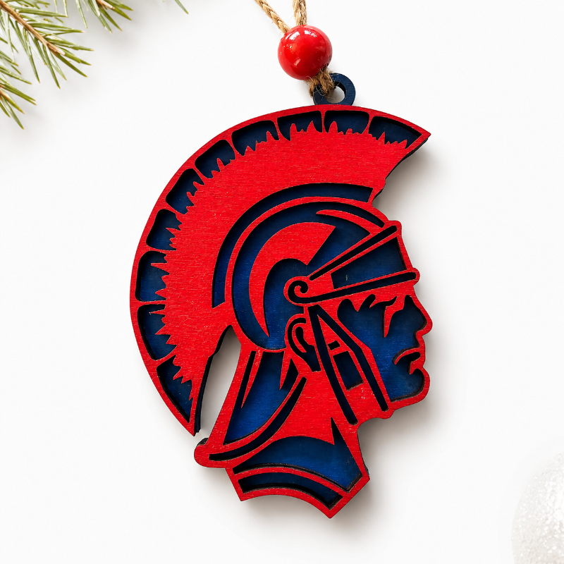 A red and navy blue wooden Spartan helmet ornament hanging from a gold string with a red bead, shown on a white background with pine branches in the corner.