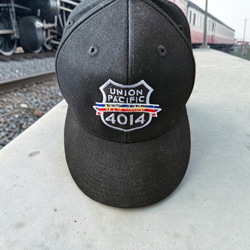 A black baseball cap with an embroidered Union Pacific 4014 logo patch, set on a concrete platform with a train and field in the background.