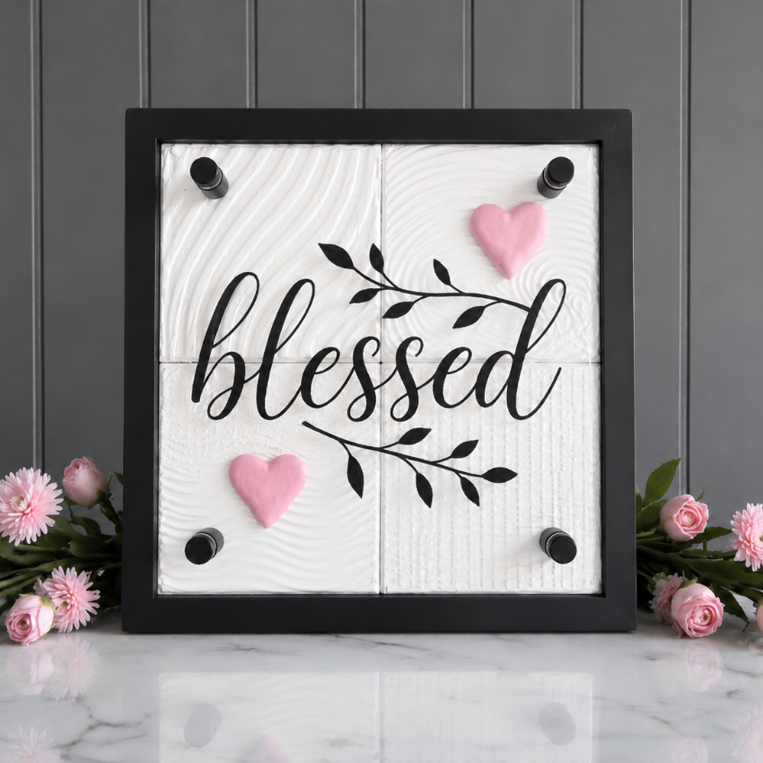 A square black-framed decorative sign with the word 'blessed' in black script on a white wood-look background, accented with two pink hearts and black branch details. The sign is displayed on a white marble surface against a gray paneled wall, with pink flowers placed on both sides.