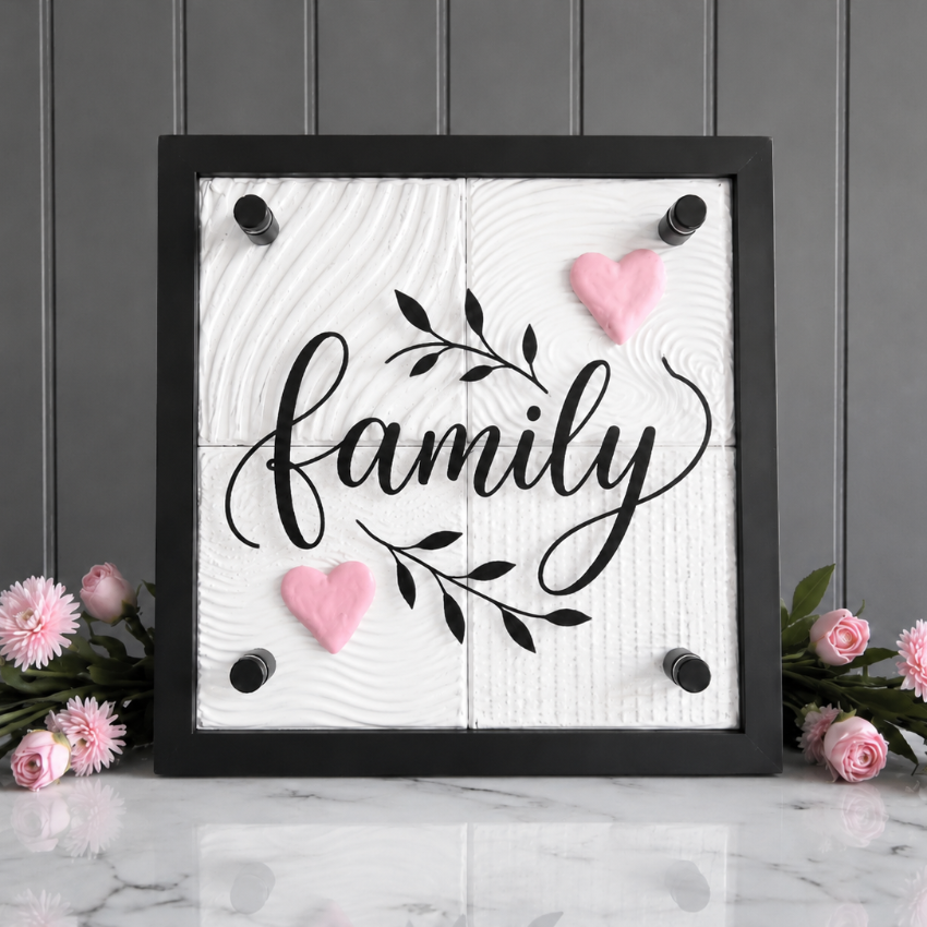 A square black-framed wall sign with the word "family" in black script over a white wood-grain background, decorated with two pink hearts and a black leafy branch. The sign is displayed on a white marble surface against a gray paneled wall, flanked by pink flowers.