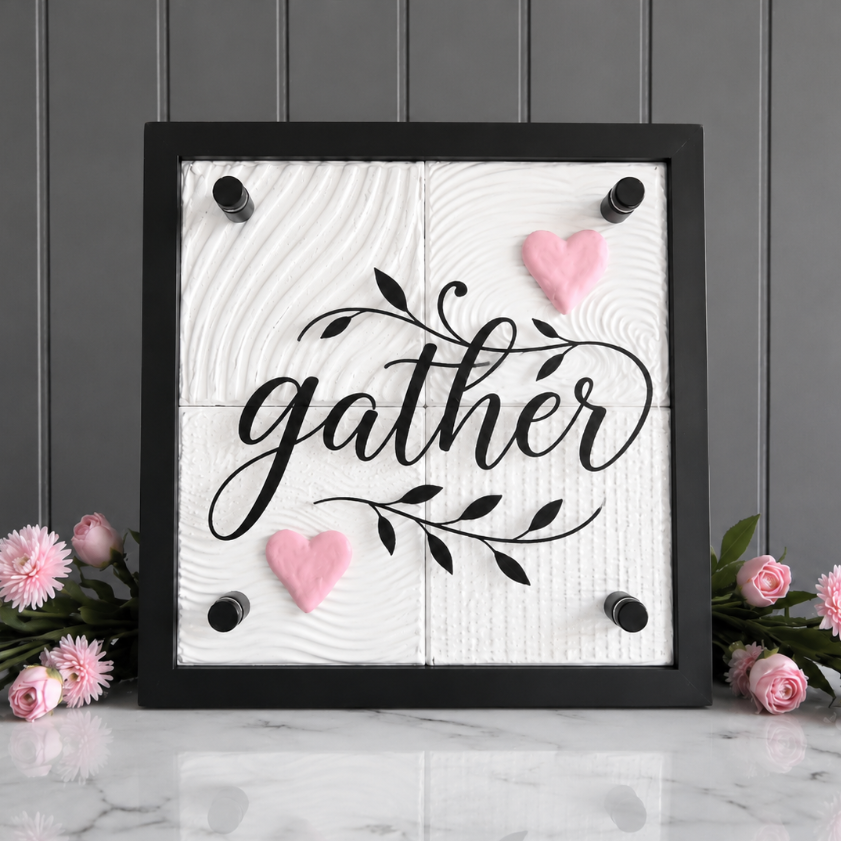 A square black-framed wall sign with the word “gather” in black script over a white wood-grain background, decorated with two pink hearts and black corner accents. The sign is displayed on a white marble surface with pink flowers on both sides against a gray paneled wall.