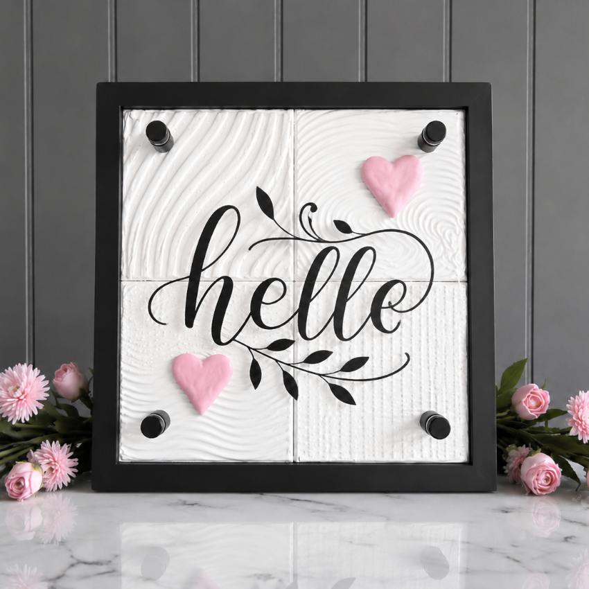 A square framed decorative sign with the word "hello" in black script over a white wood-grain background, accented with two pink hearts and black corner knobs. The sign is displayed on a white marble surface against a gray paneled wall, flanked by pink flowers on both sides.