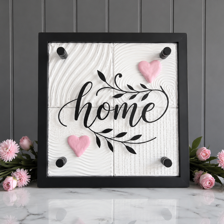 A square black-framed decorative wall sign with the word "home" in black script over a white wood-grain background, accented with two pink hearts and black corner knobs. The sign is displayed on a white marble surface with pink flowers against a gray paneled wall.