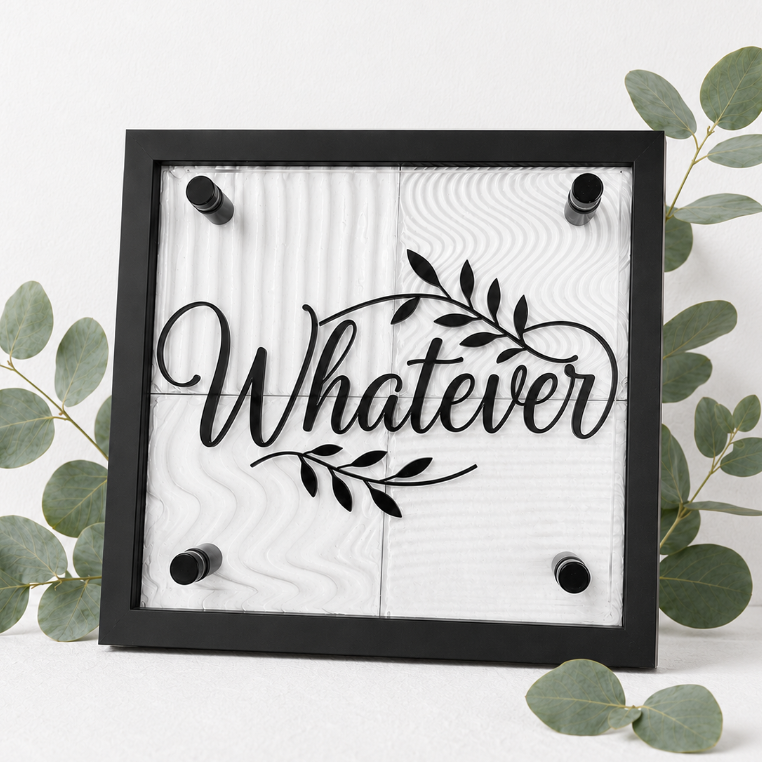 A square black-framed wall sign with the word 'Whatever' in black script over a white wood-grain background, styled with eucalyptus branches on a white surface.